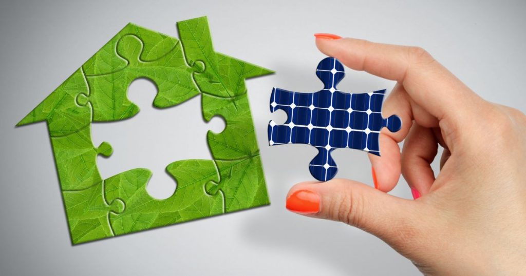Een dame houdt een puzzelstukje met zonnepaneelpatroon boven een puzzel met groene puzzelstukken in de vorm van een huisje.