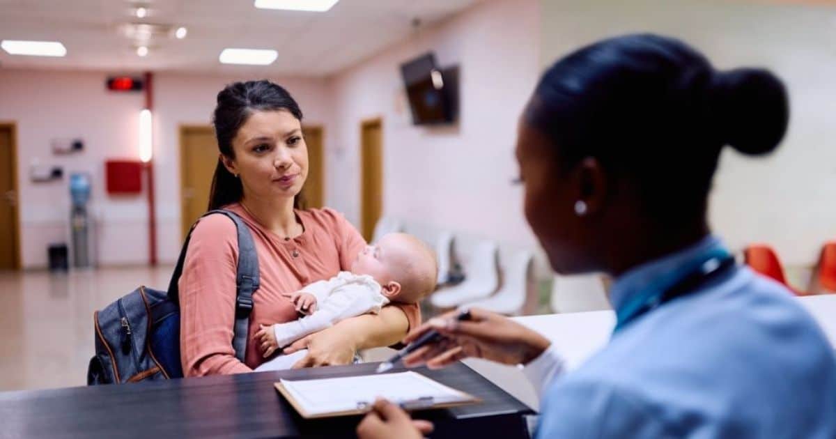 Een moeder met zwart haar, een roze hemd en een rugzak staat met haar baby in de armen voor de balie in een ziekenhuis. De baby is in het wit gekleed. Achter de balie zit een verpleegster met zwart haar in een dot. De verpleegster geeft de moeder een klembord met papier en een pen.