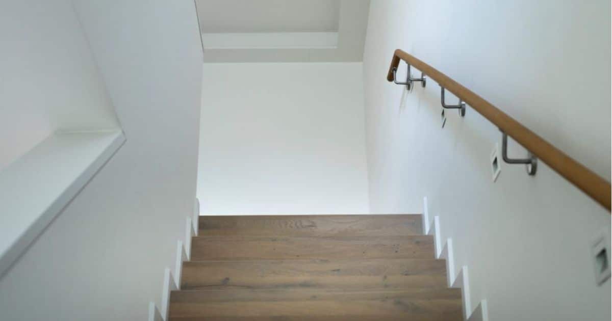 Een molenaarstrap met een leuning die aan een witte muur hangt in een voor de rest volledig witte ruimte.