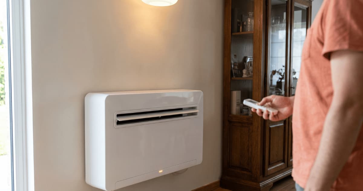 Een persoon in een roze top richt een afstandsbediening op een witte airco vloerunit laag tegen de muur in een woonkamer met een houten kast ernaast.