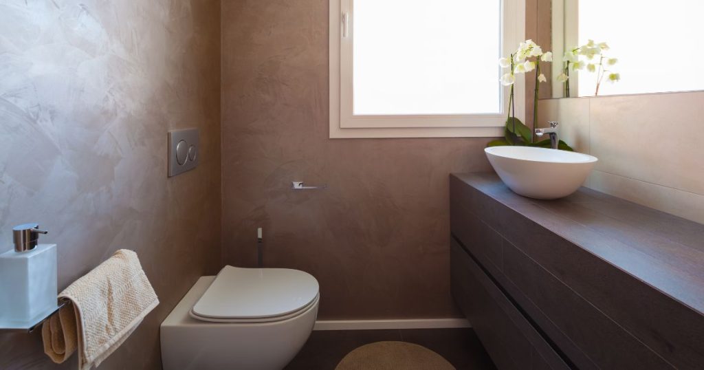 Compacte mortex badkamer met bruine wanden, houten meubel en ronde waskom onder raam.