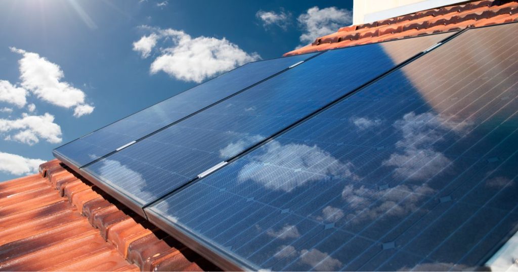Drie zonnepanelen op een dak met pannen weerspiegelen de blauwe hemel en witte wolkjes boven hen.