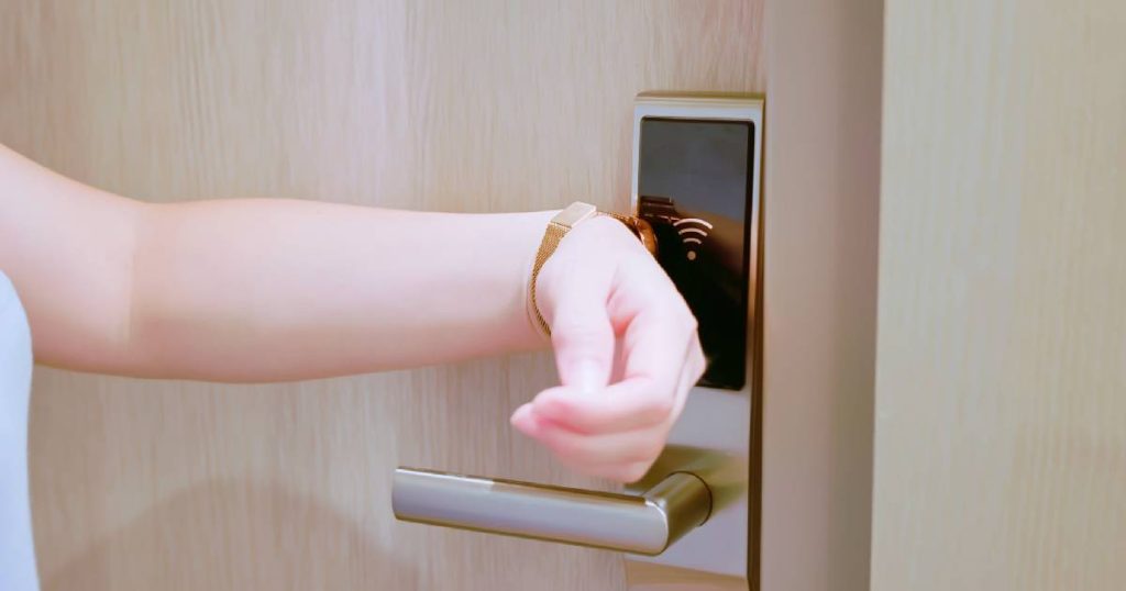 Een hotelgast opent de deur van haar hotelkamer met de NFC-tag in haar polsbandje. De eikenhouten deur heeft een metalen klink waarin een NFC-lezer zit verwerkt.