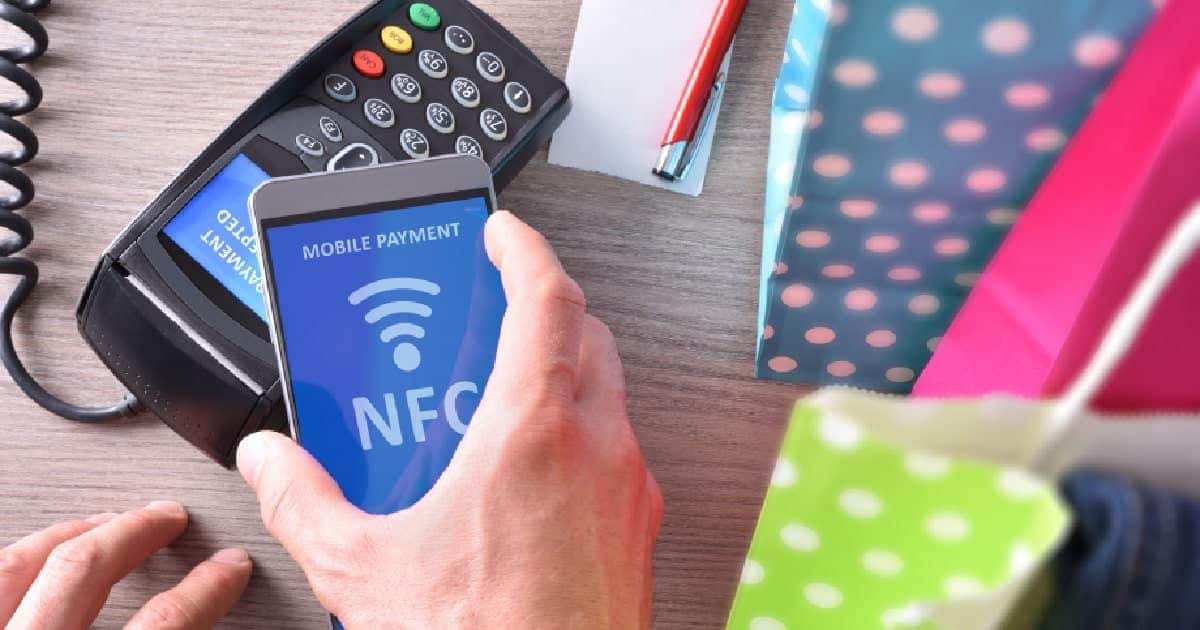 Hand houdt een smartphone met "Mobile Payment" en het NFC-symbool op het scherm tegen een betaalterminal, met kleurrijke boodschappentassen op de achtergrond.