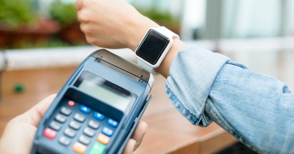 Een klant met een lichtblauw hemd gebruikt haar wearable om een transactie af te ronden met een mobiele, NFC-ondersteunende betaalterminal