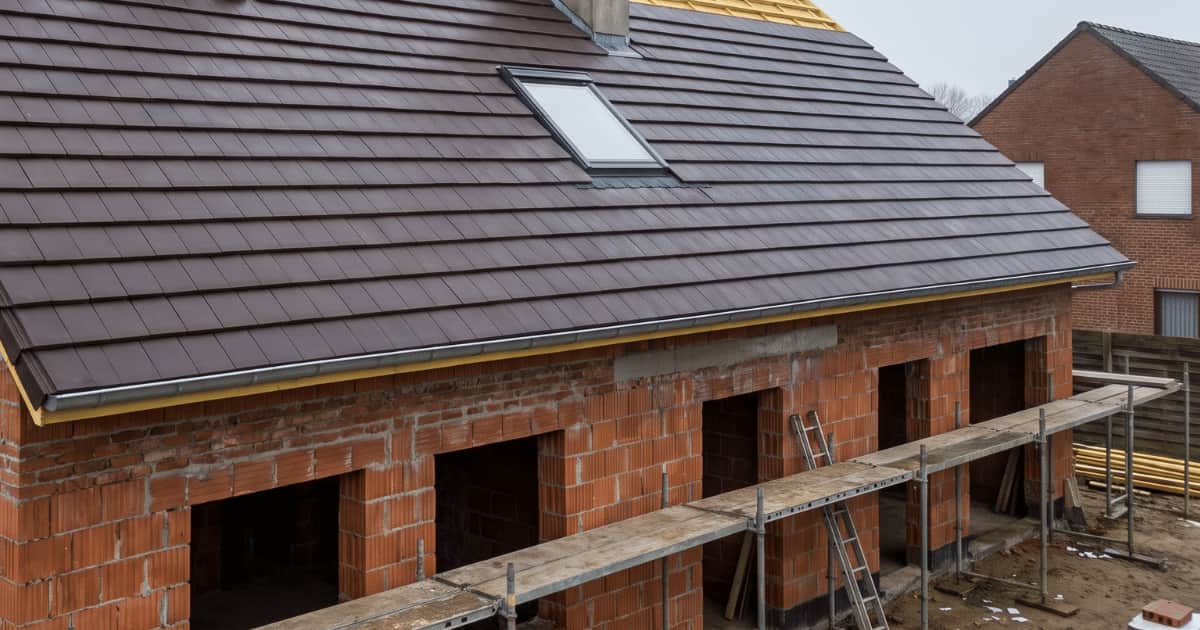 Een nieuwbouwwoning in ruwbouw toont een hellend dak bedekt met donkerbruine vlakke dakpannen met een ingebouwd dakraam terwijl de rode snelbouwstenen gevelmuren met open raamopeningen en steigerwerk eronder zichtbaar zijn onder een bewolkte lucht.