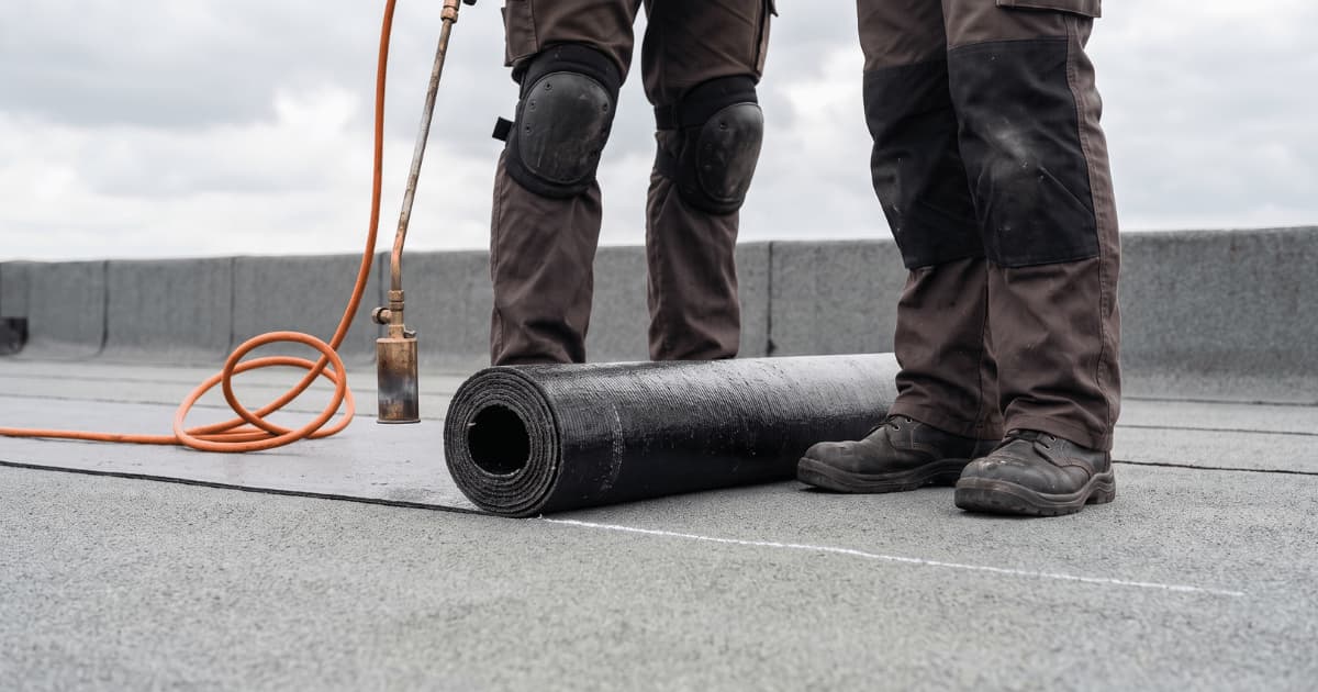 Een dakwerker staat op een plat dak en brandt met een gasbrander een donkere bitumenstrook vast op de ondergrond.