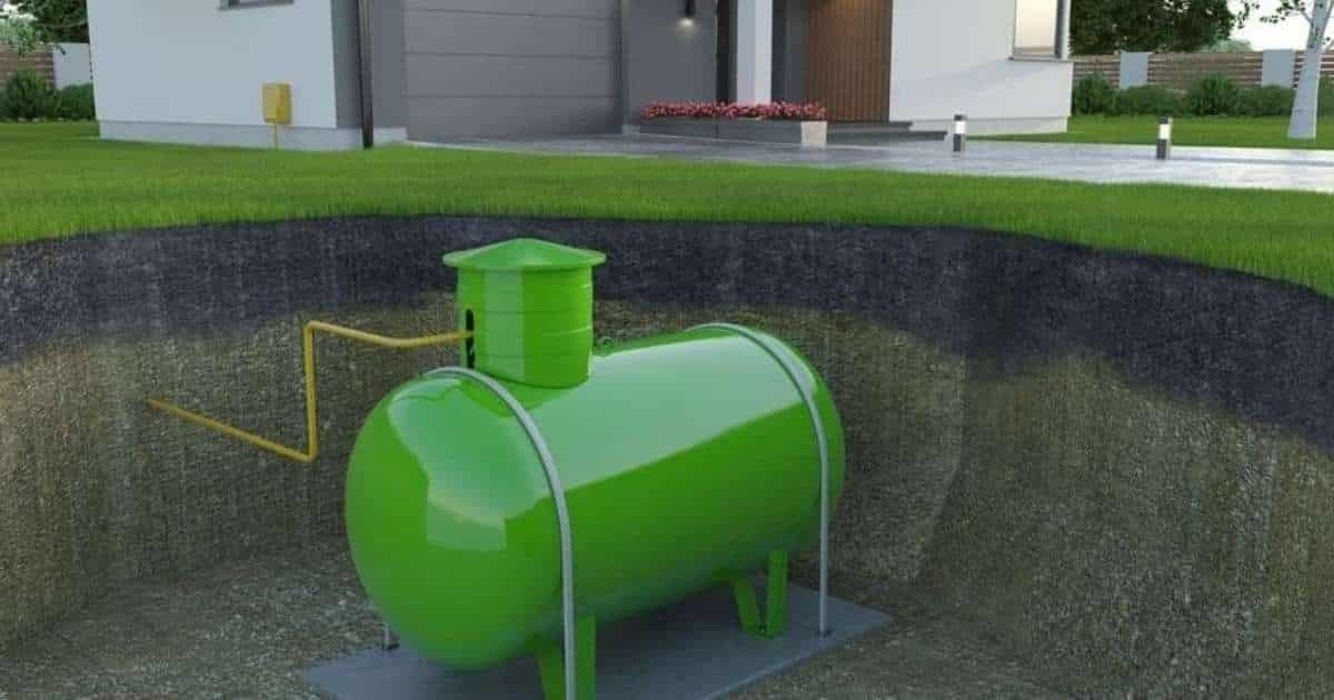 Een groene gastank, ingegraven onder de grond in de tuin rond een moderne witte woning met grijze garagepoort, donkerhouten deur en lange oprit met verlichting. Een gele slang verbindt de gastank met de woning.