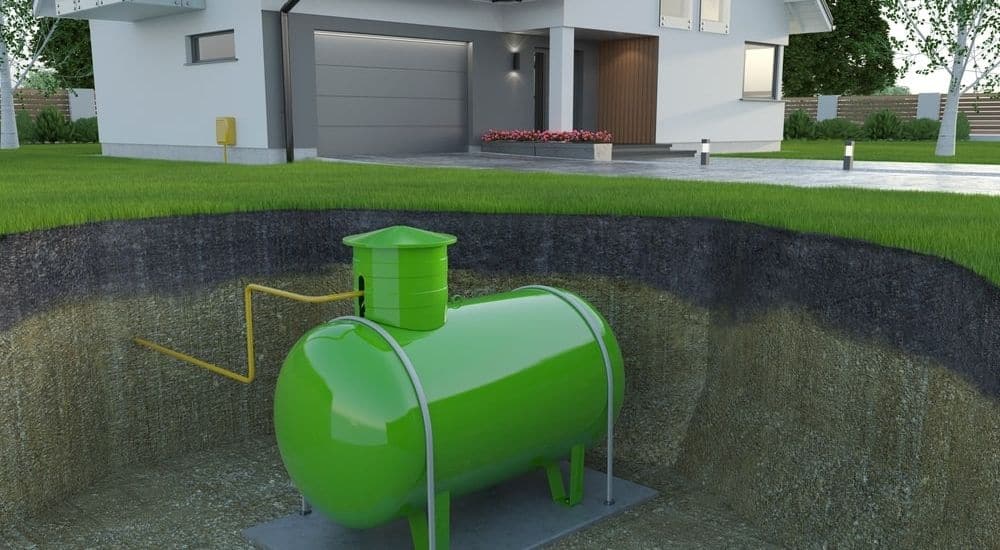 Een groene gastank, ingegraven onder de grond in de tuin rond een moderne witte woning met grijze garagepoort en lange oprit.