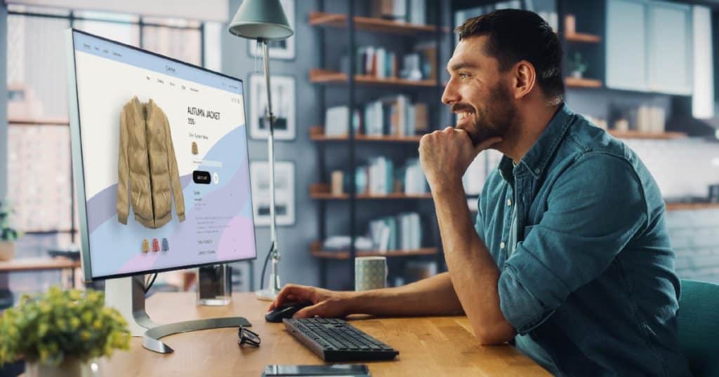 Een man in een blauw jeanshemd kijkt lachend naar de moderne monitor van zijn computer. Op het beeld staat een webshop waar een beige jas verkocht wordt.