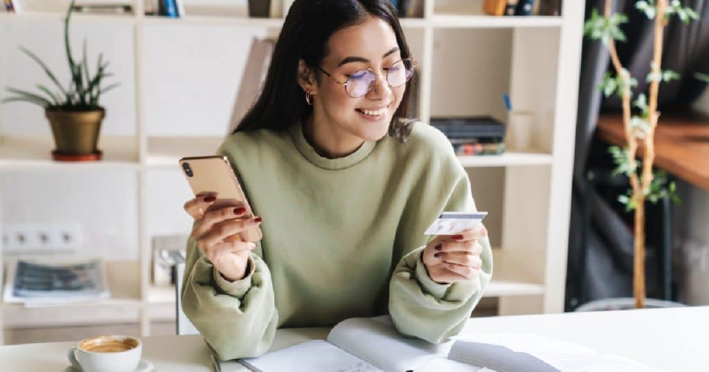 Een vrolijke dame met een ronde bril en een groene trui gebruikt haar kredietkaart of debetkaart om een online aankoop te doen met haar smartphone.
