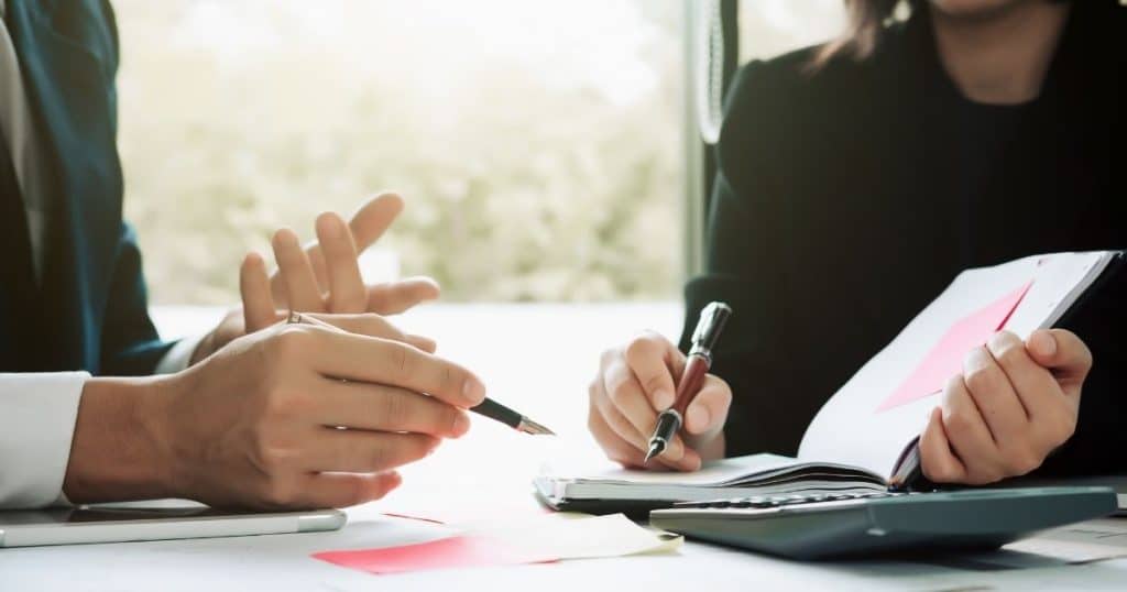 Een man en vrouw zitten in formele kledij aan een witte tafel. Op de tafel liggen documenten en een rekentoestel. Ze houden beide een pen vast.