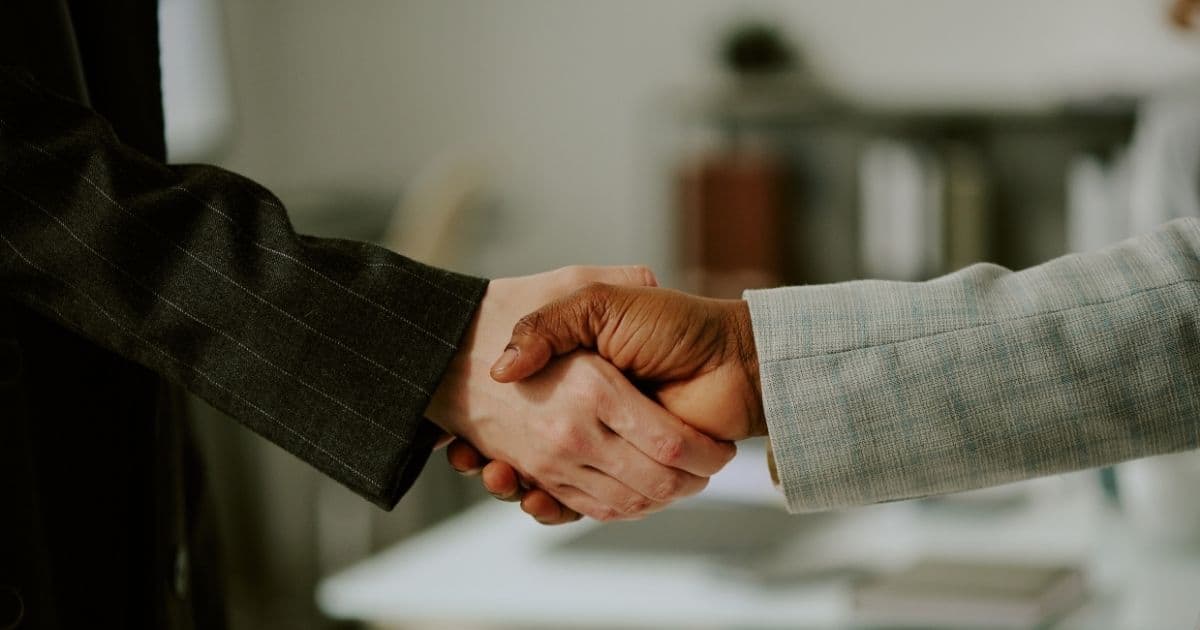 Twee mensen schudden elkaar de hand. Enkel hun armen zijn in beeld. De linkse draagt een zwarte blazer met witte krijtstreep. De rechtse een groene blazer met licht ruitjespatroon.
