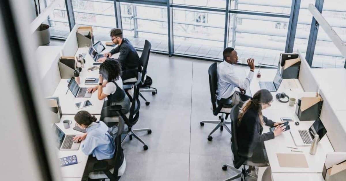 Een open kantoorlandschap van bovenaf bekeken: medewerkers werken op hun laptop(s), drinken van een fles water, of scrollen achteloos op hun smartphone of tablet.