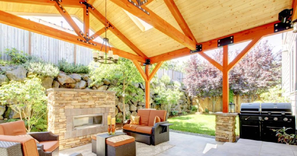 Een moderne open eiken serre. Onder de serre staan een zwarte barbecue, enkele leren stoelen en een stenen haardvuur.