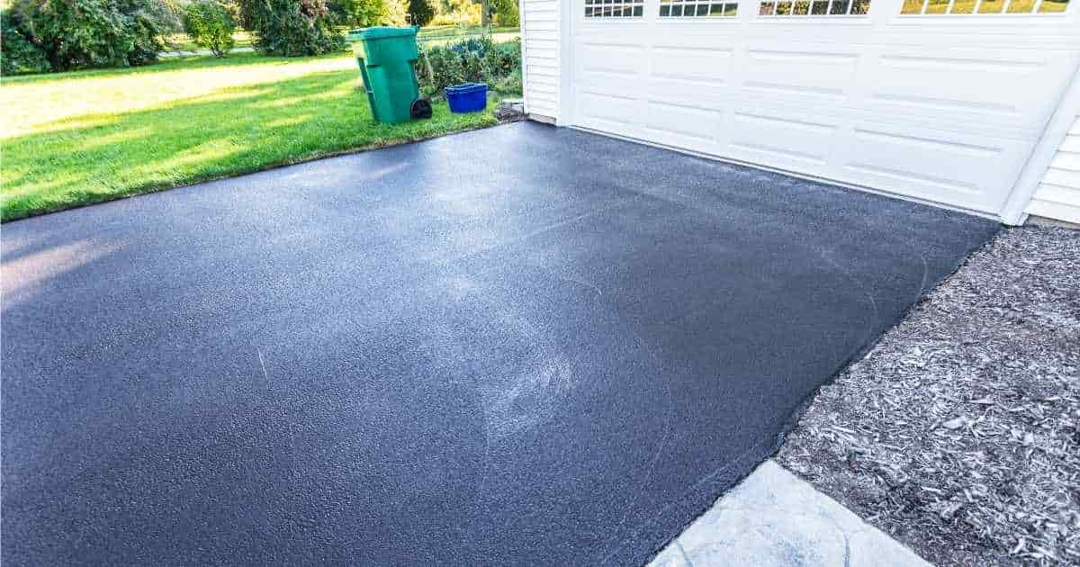 Een oprit uit asfalt geeft uit op een witte garagepoort. Naast de oprit ligt een grasveld, waarop een groene vuilnisbak staat.