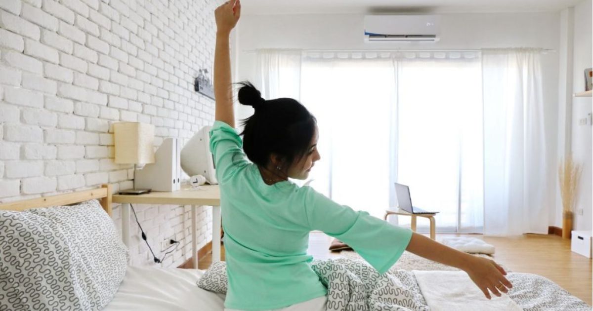 Vrouw die zich uitstrekt op bed in een lichte slaapkamer met een airco aan de muur.