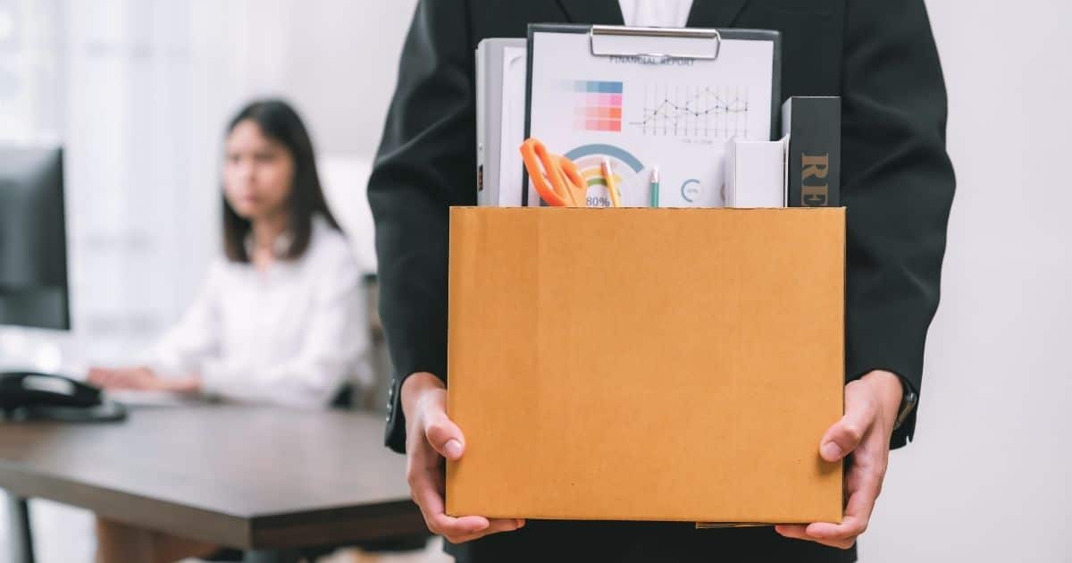 Het bovenlichaam van een man in zwart kostuum. Hij heeft een grote kartonnen doos vast met daarin een klembord, schaar, potloden en boeken: hij heeft net zijn ontslag gekregen. Op de achtergrond zit een ex-collega aan haar bureau achter een computerscherm. Ze heeft donkerbruin haar en draagt een wit hemd.