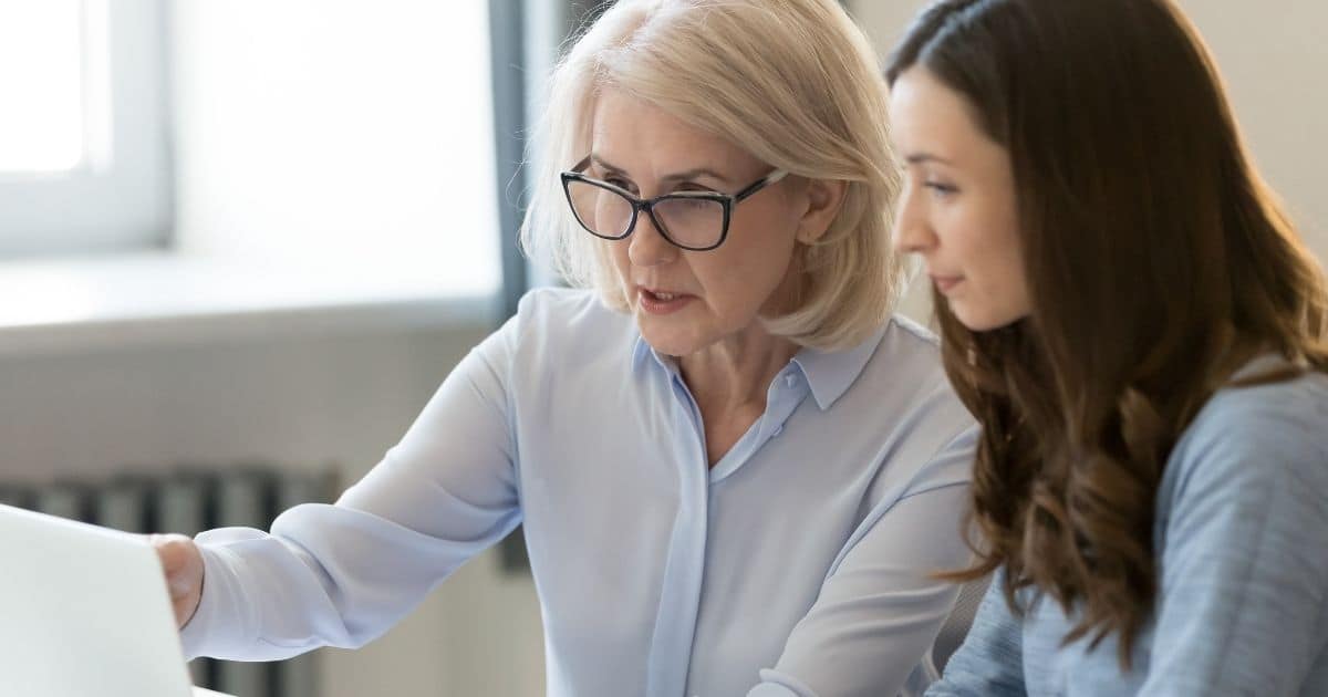 Een werknemer met wit haar, blauwe bril en lichtblauw hemd wijst naar een computer. Ze toont iets aan haar jongere collega: een dame met bruin haar en een blauw hemd.