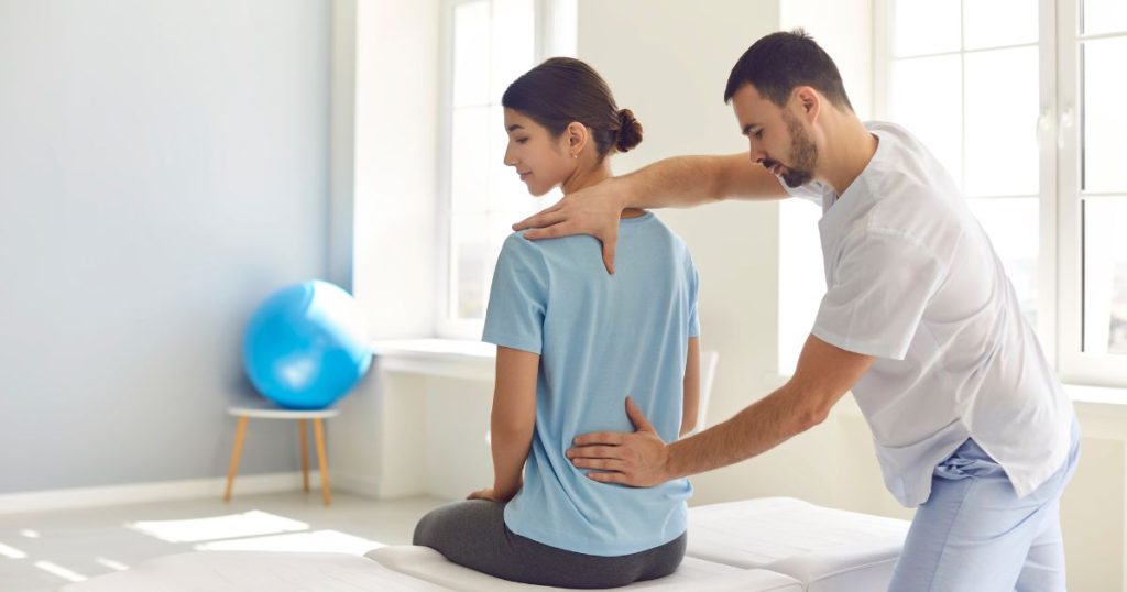 Een man met baard en wit hemd houdt zijn handen tegen de rug van een dame die op een bank zit. De twee duimen van de osteopaat wijzen naar elkaar.