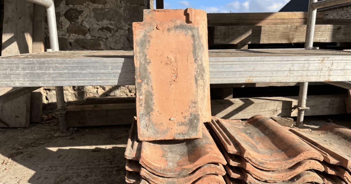 Een verweerde terracotta Boomse pan staat rechtop op een stapel gelijkaardige recuperatiepannen en toont zijn vlakke onderzijde met het neusje bovenaan terwijl aluminium stellingbuizen en houten balken op een bouwwerf de achtergrond vormen in fel zonlicht.