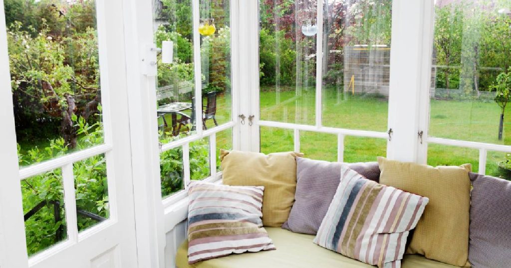 Een oude veranda die na het renoveren beschikt over nieuwe, dubbele beglazing. In de veranda staat een zetel met kussens. De ramen geven uit op een knusse, groene tuin.
