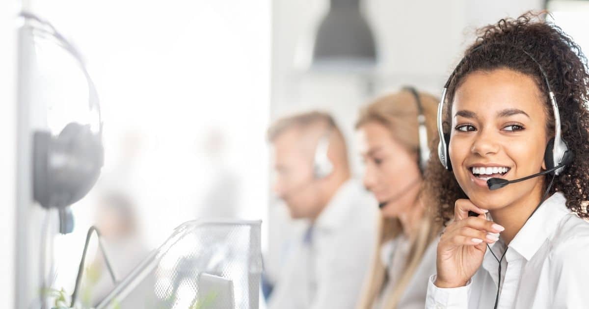 Een dame met wit hemd en een head set. Op de achtergrond een dame en heer, eveneens in wit hemd en met headset. Ze zitten achter computers.