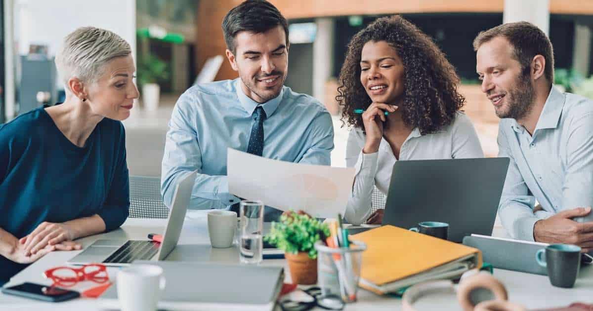 Een team callcenterspecialisten zit rond een tafel met computers, mappen en papieren. Ze bespreken een marketingstrategie.