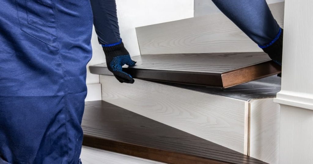 Man in blauwe werkbroek en met donkere handschoenen vervangt de overzettreden tijdens een traprenovatie. 