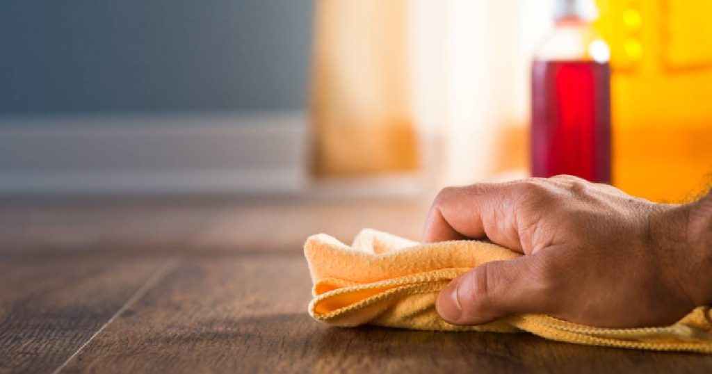 Een hand brengt olie aan op een parketvloer met een oranjegele microvezeldoek.