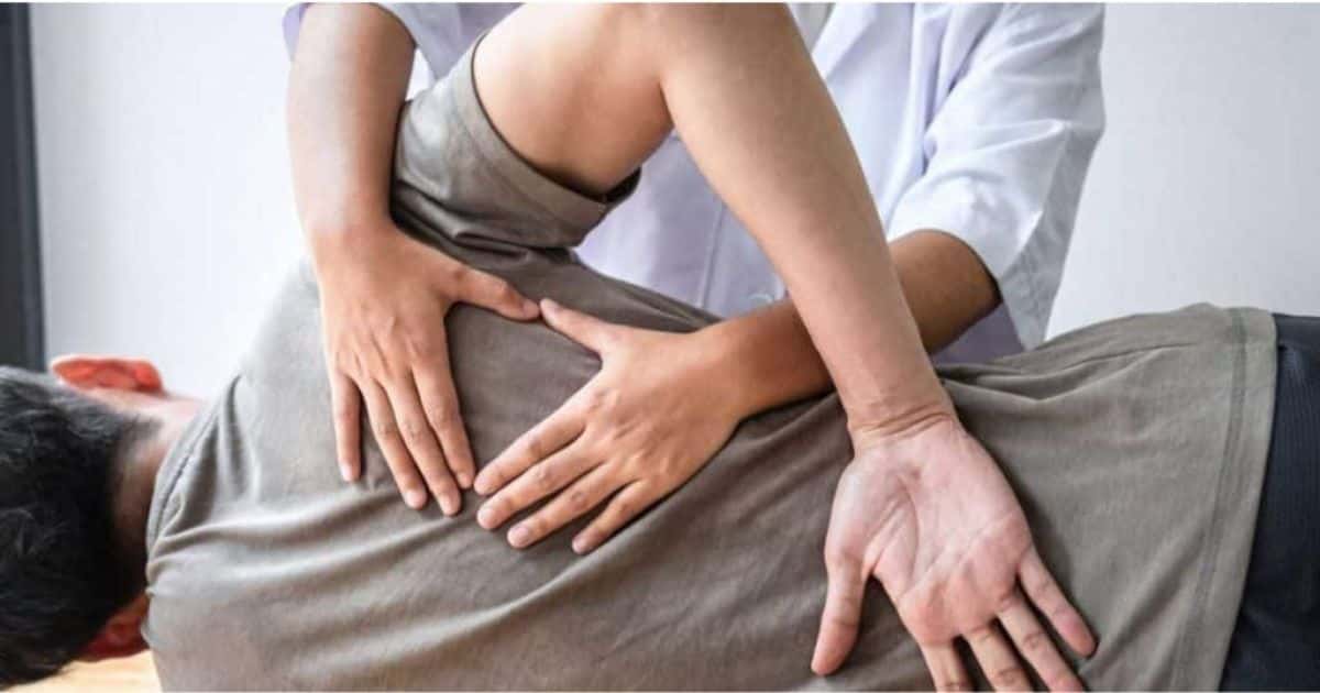 Een patiënt met rugklachten wordt behandeld door een kinesist. De patiënt houdt één hand open op zijn rug. De kinesist houdt beide handen net onder de schouder.