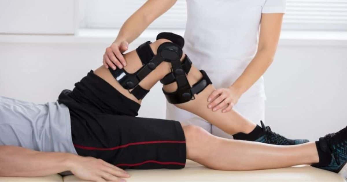 Een patiënt met knieproblemen bij de kinesist: rond zijn knie is een speciale beugel. De kiné houdt haar handen op het boven- en onderbeen.