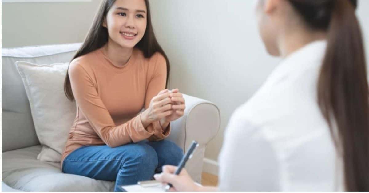 Een jonge patiënte zit in een beige zetel voor een consultatie bij een vrouwelijke psychiater.