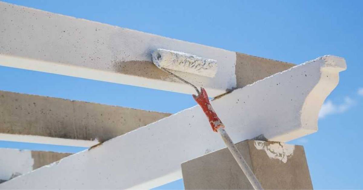 Iemand schildert met een verfroller op een steel de betonnen balken van een pergola wit.