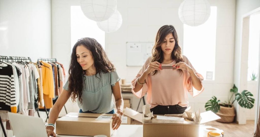 2 dames staan naast elkaar in een grote, witte ruimte. De dame met blauw-groene blousse richt haar rechterhand naar een computerscherm, terwijl ze met de linkerhand een doos vasthoudt. De dame met roze blousse neemt ondertussen een foto met haar smartphone.