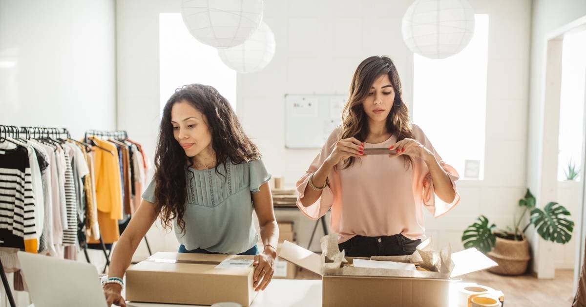 Een werkgever en haar eerste werknemer werken samen in een kleine kledingzaak. Ze verpakken kledij om te verzenden naar klanten die online een bestelling plaatsten. De linkse dame (met groen t-shirt en bruin, krullend haar) gebruikt haar laptop om de verzending te registreren. De rechtse dame (met roze t-shirt en bruin golvend haar) fotografeert de inhoud van een pakket met haar smartphone. Op de achtergrond staan links kledingrekken, rechts een kamerplant in een grote pot.