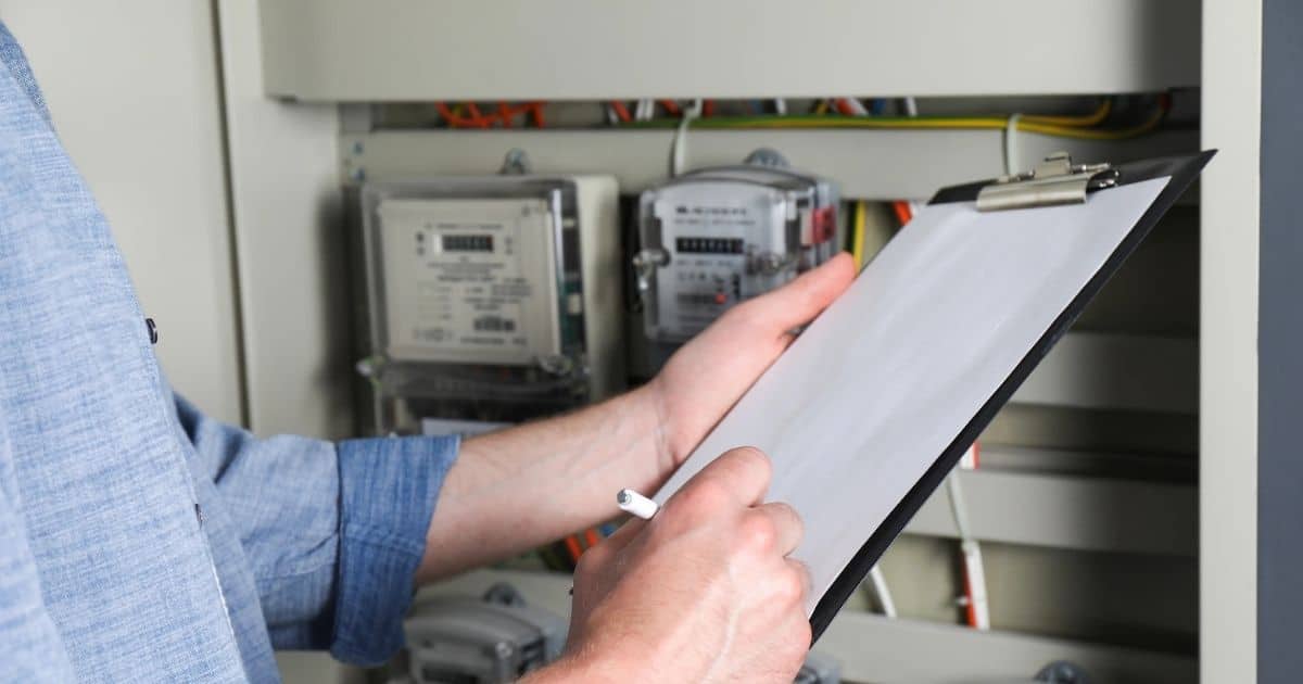 Een persoon houdt een klemblok met leeg blad voor een meterkast. Hij of zij schrijft iets op het blad, en draagt een lichtblauw hemd.