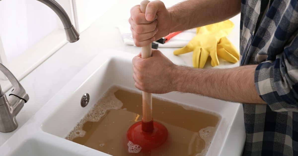 Een man met geruit hemd gebruikt een zuignap of plopper om een verstopte gootsteen te ontstoppen. De gootsteen is wit, maar er staat een laag troebel bruinoranje water met schuim in.