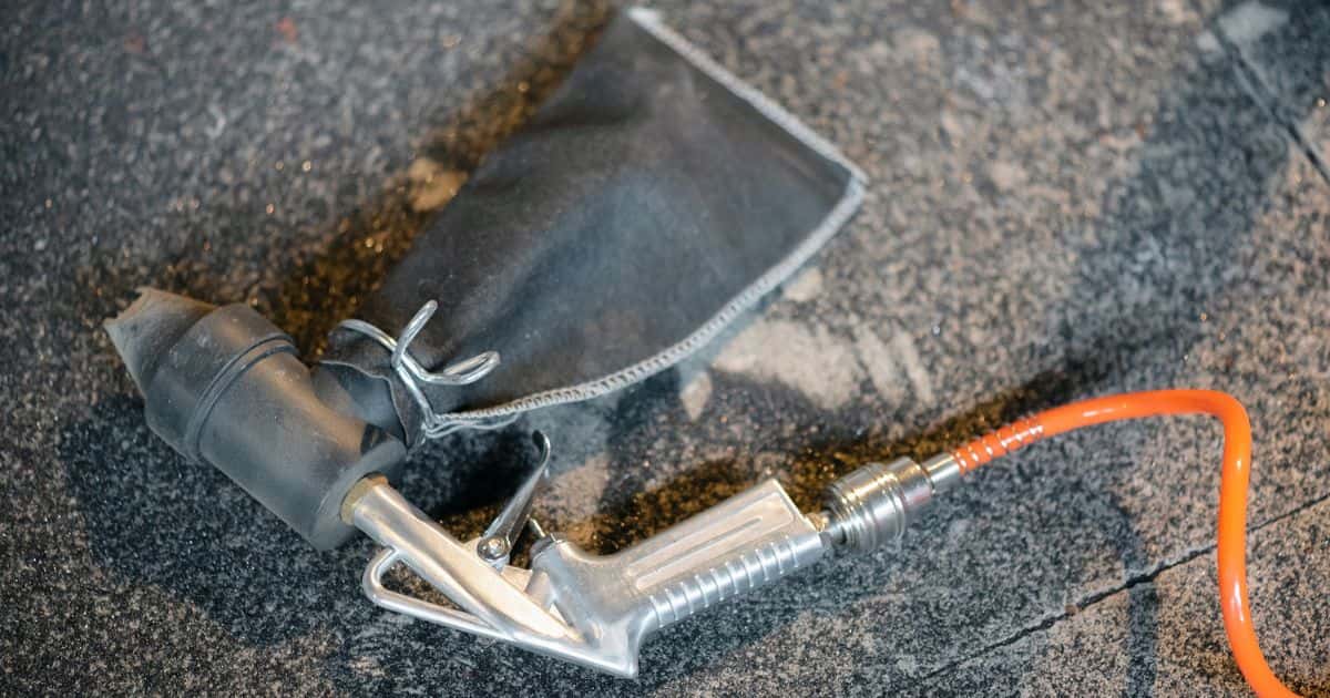 Detailbeeld van een zandstraalpistool: inox of chroom pistool met opzetstuk in kunststof en stoffen zakje. Het pistool wordt aangedreven via een oranje buisje.