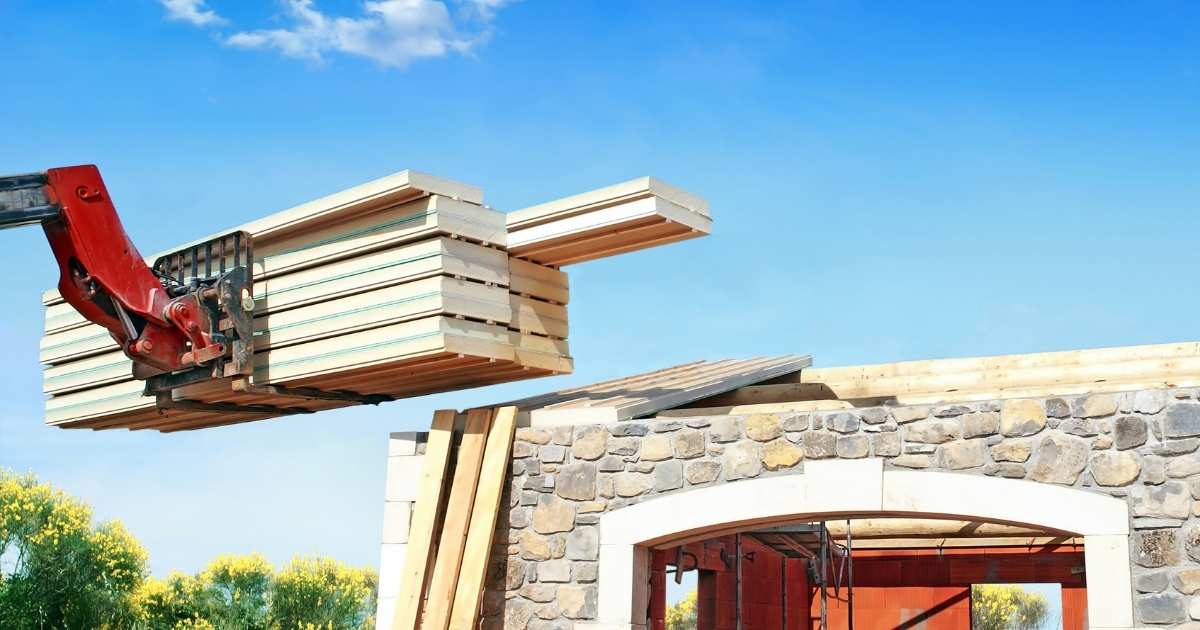 Een kraan tilt een stapel sandwichpanelen op een hellend dak. Het dak ligt bloot: enkel het houten dakgebinte is zichtbaar.