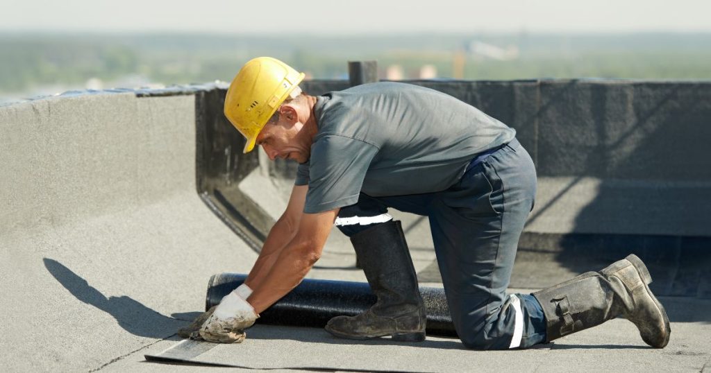Een dakwerker legt nieuwe dakbedekking op een plat dak. Hij draagt een gele veiligheidshelm, grijs werkshirt, blauwe werkbroek en zwarte laarzen.
