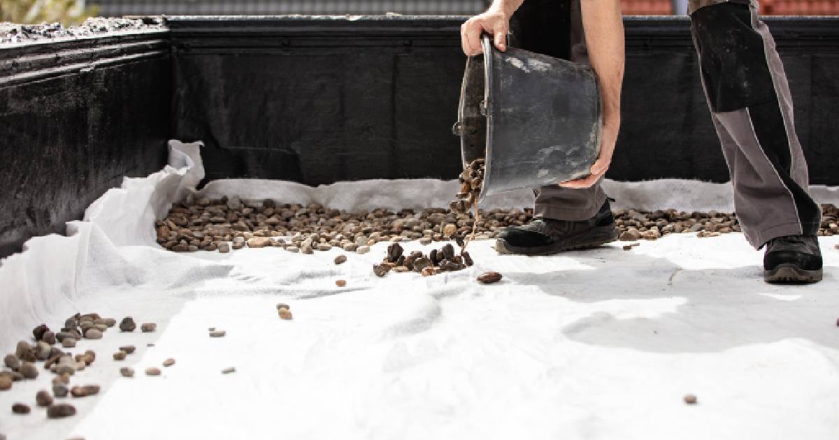 Een dakwerker werkt een plat dak af met grind na het dak te herstellen. Hij giet het grind uit een emmer op de bovenste laag dakafdichting.