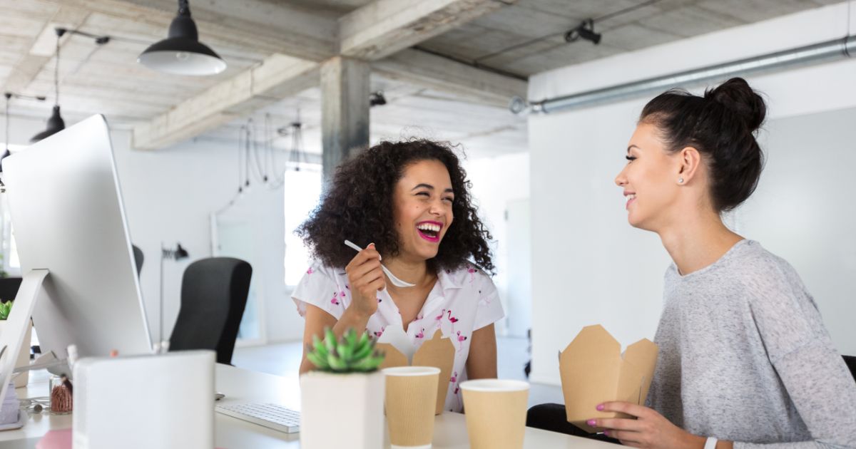 Twee vrouwelijke collega's eten een afhaalmaaltijd aan hun bureau.