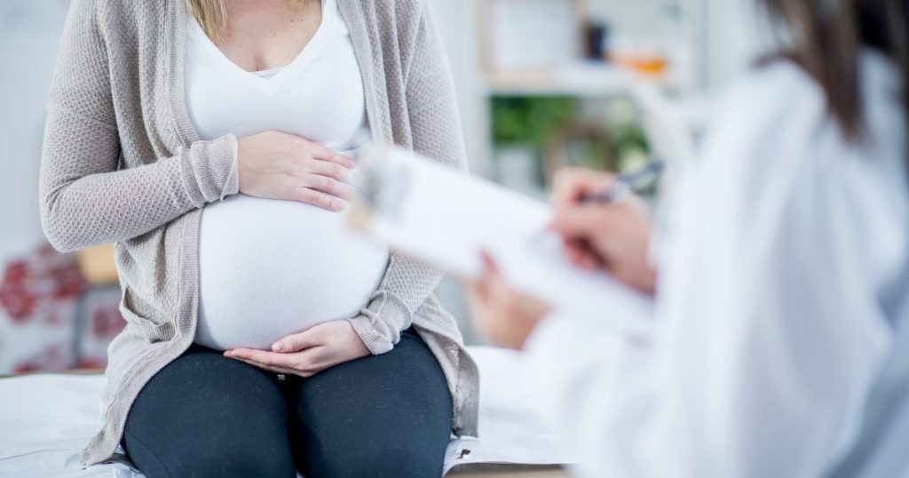 Een zwangere vrouw in wit t-shirt en grijs vest bij de dokter.