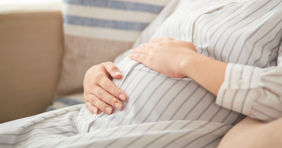 Een zwangere vrouw in gestreepte jurk op een beige zetel. Ze houdt de handen op haar buik om de baby te kunnen voelen.