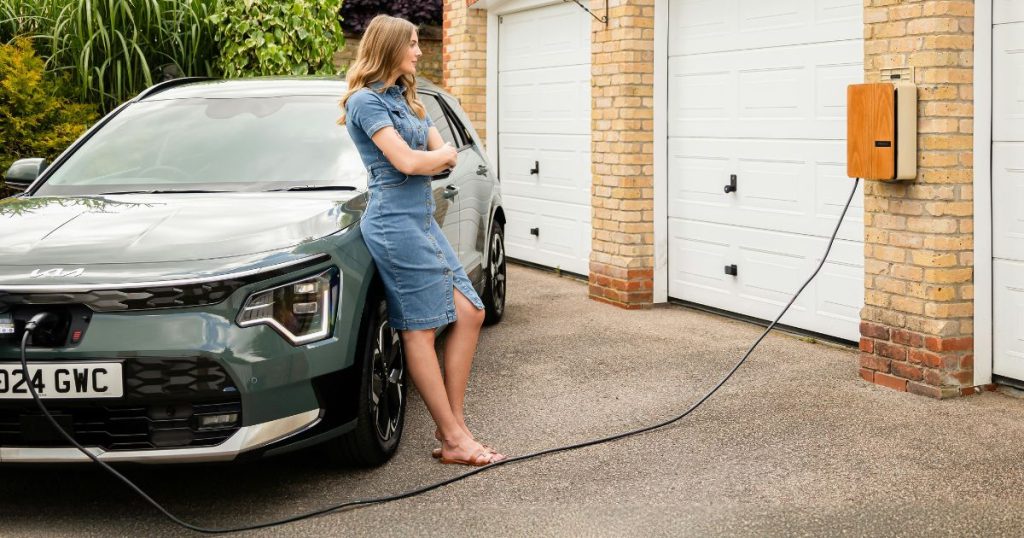 Een dame in jeansjurk leunt tegen haar grijsgroene elektrische wagen terwijl ze wacht tot de auto is opgeladen. De auto is via een laadkabel verbonden met de laadpaal, die gemonteerd is tegen de oranje bakstenen muur tussen verschillende garageboxen met witte garagepoorten.
