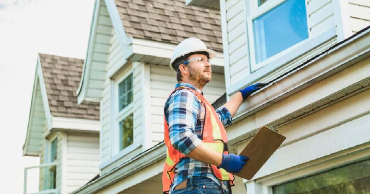 Een dakspecialist inspecteert de huidige staat van een dak met dakpannen en verschillende dakkapellen om te bepalen wat de prijs van een nieuw dak voor dit huis zou bedragen. Hij draagt een witte veiligheidshelm, blauwe handschoenen, een blauw met wit geruit hemd, een bril en een oranje met geel fluohesje. In zijn hand heeft hij een notitieblok vast.