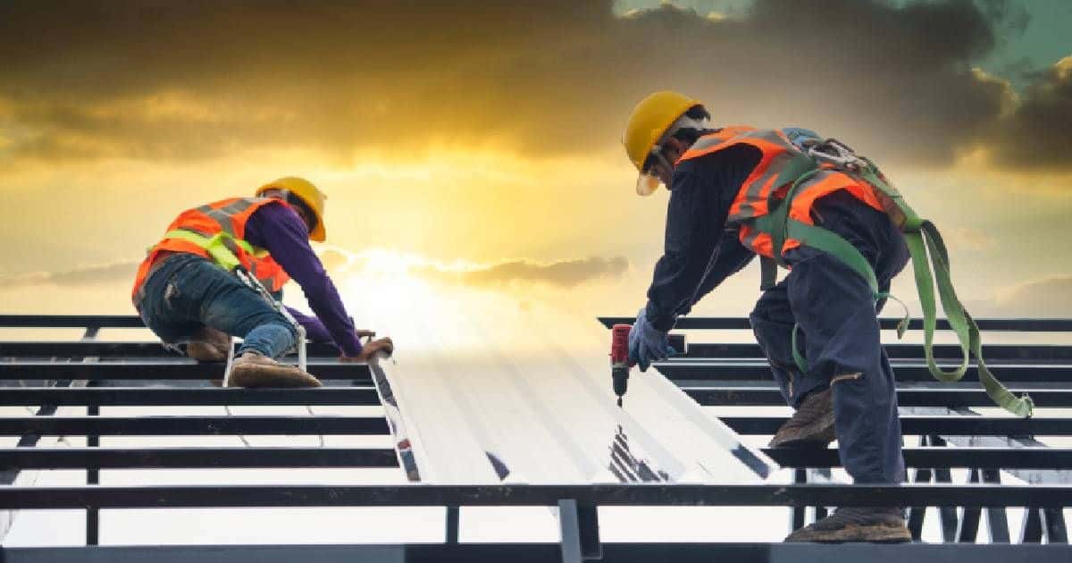 Twee dakwerkers bevestigen dakplaten op een nieuw dak, met op de achtergrond de ondergaande zon. De dakwerkers dragen gele veiligheidshelmen, brillen, fluohesjes en een blauwe overall. 