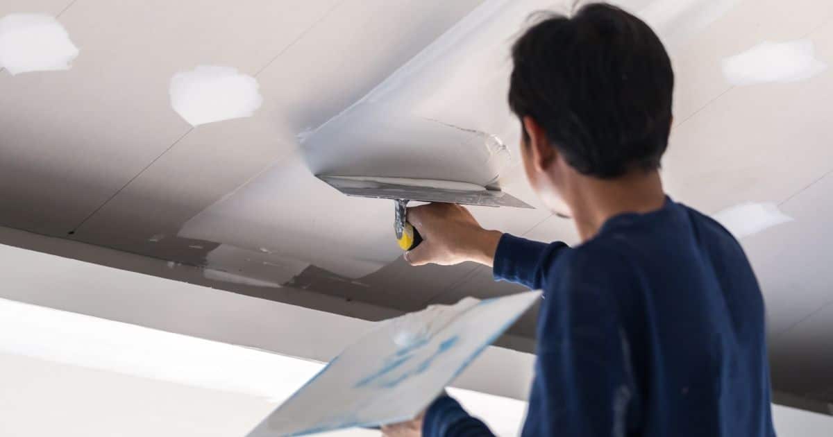 Een stukadoor of plakker brengt pleister aan op een plafond. Hij draagt een blauwe trui en gebruikt een raapbord en spaan.