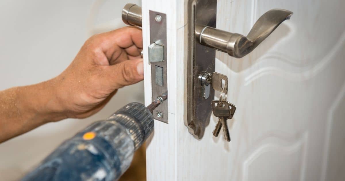 Een slotenmaker gebruikt een boor om een nieuw slot vast te maken in de witte houten deur van een appartement. De sleutels zitten in het slot.